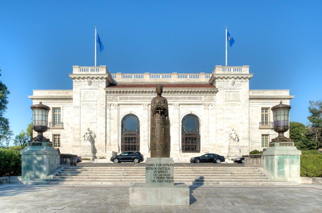 Monumental white marble building with triple-arched entrance behind bronze statue of a woman