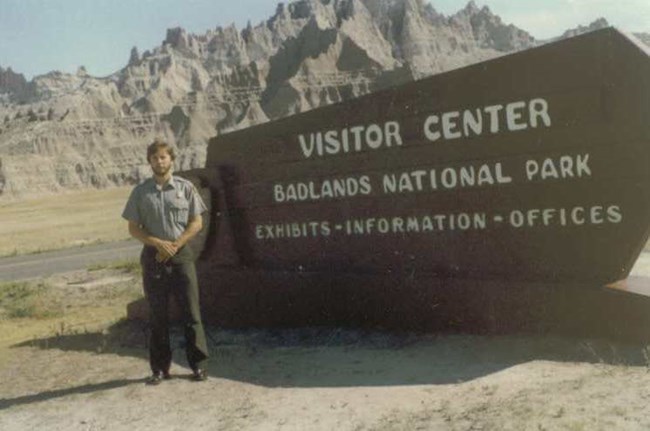 A young man with a brown beard and park ranger uniform poses with the Visitor Center sign for Badlands National Park. In the background are eroded sedimentary features and grass.