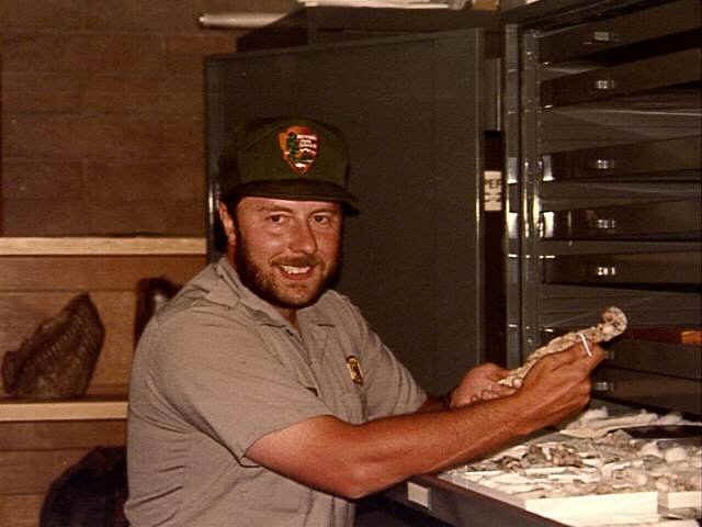 Photograph of a man in a ranger uniform sitting in front of a museum cabinet, holding a fossil specimen.