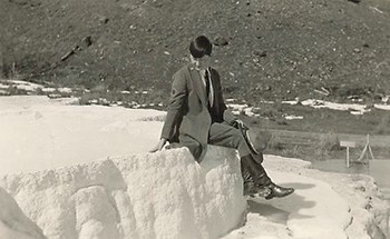 Marguerite Lindsley wearing a ranger uniform and holding a stetson hat sitting down on a natural feature with her ankles crossed.