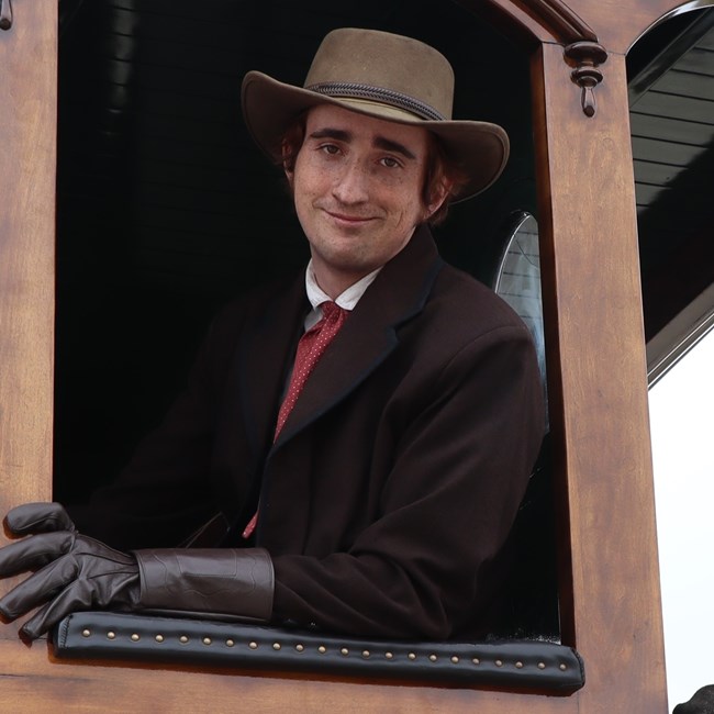 A close up shot of a young man wearing a hat and gloves smiles from the window of a steam locomotive.