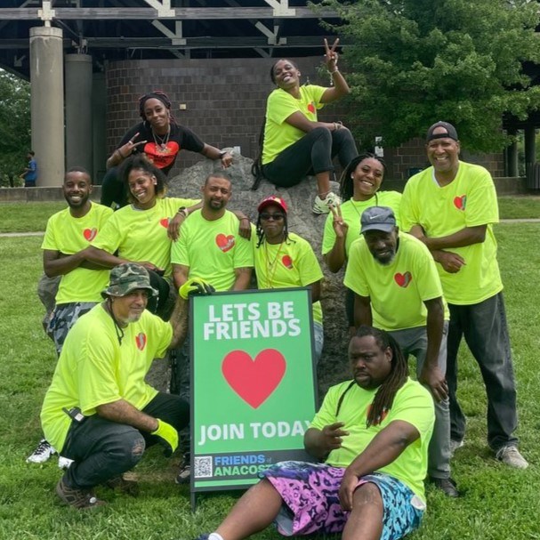 A group of 11 people wearing matching neon yellow t-shirts with hearts smiling alongside a sign that says ‘Lets Be Friends – Join Today.’