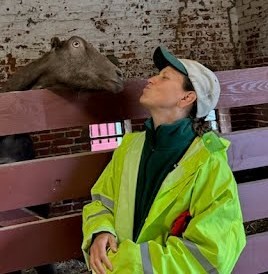 A woman wearing a neon yellow jacket makes a kissing face towards a goat in a pen.