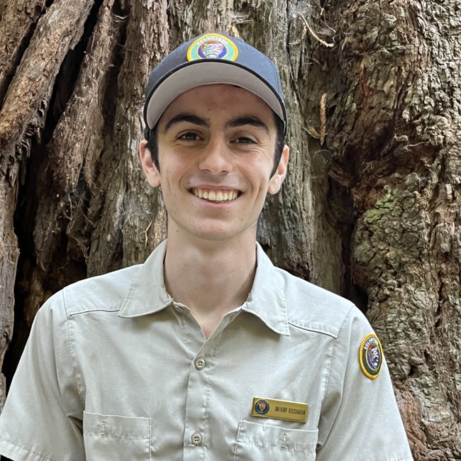 A young man wearing a volunteer hat and shirt smiling in front of tree bark.