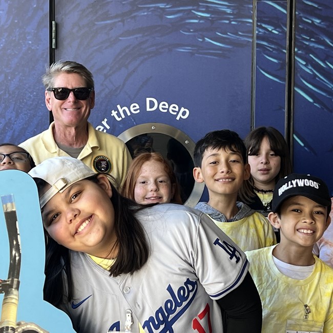 A man in a yellow polo shirt and sunglasses smiling alongside eight young students.