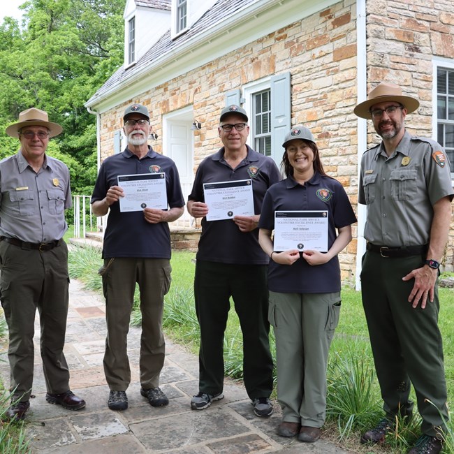 Three adults wearing volunteer polos and hats hold certificates, smiling in between two smiling rangers.