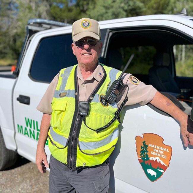 A man wearing a volunteer hat, vest, and polo smiles in front of a National Park Service truck with a walkie talkie.