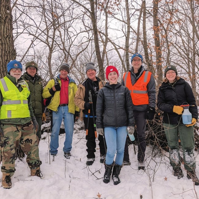 Six adults wearing various VIP and winter gear smile alongside a park ranger in a snowy terrain, holding loppers.