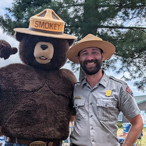 A ranger in uniform smiles with a mascot of Smokey the Bear.