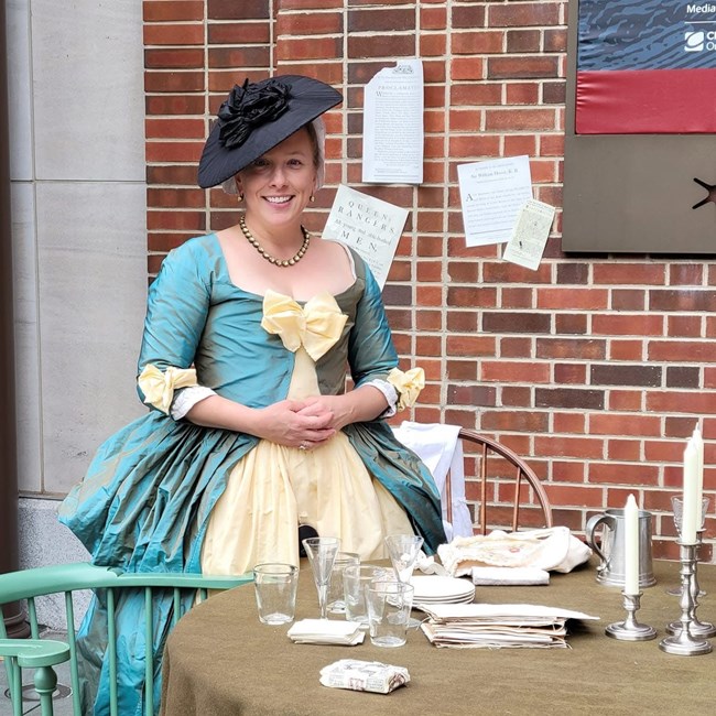 A woman wearing a revolutionary style hat and dress in behind a table with glasses and candlesticks.