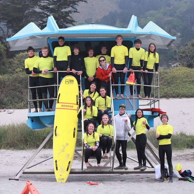 A group of 16 young adults wearing yellow t-shirts and wet suits smile on a lifeguard stand alongside two NPS staff members.