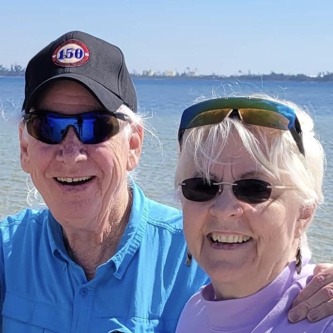 A man in a hat and sunglasses smiling alongside a woman in sunglasses in front of a waterfront.