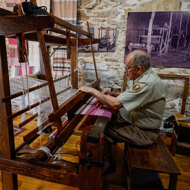 A man in a volunteer polo sitting at a large loom, weaving a pink textile.