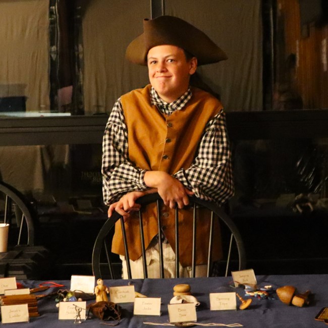 A young man wearing colonial dress stands behind a table with various colonial toys and games.