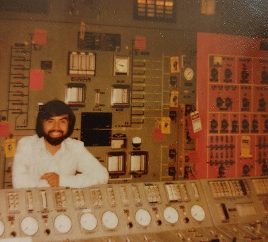 Young man with a full beard and black hair stands between a bank of industrial gauges and in front of a wall with more gauges, dials, and controls.