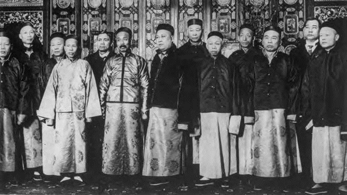 Black and white photo of a group of men in Chinese dress with ornate wallpaper in the background.