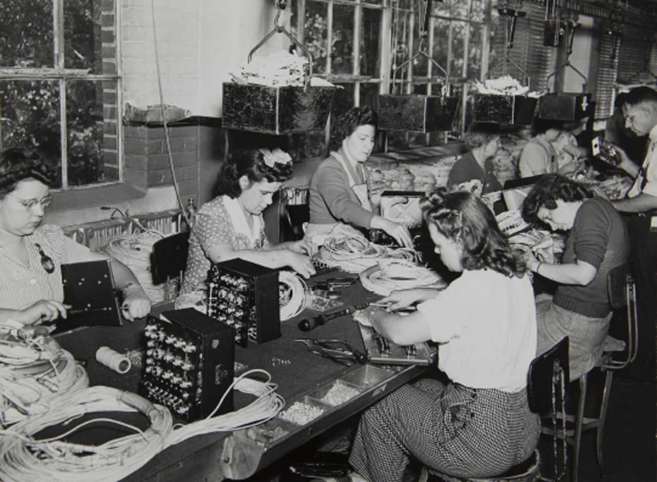 women sitting at long table facing each other and working on wiring parts for gun electonics