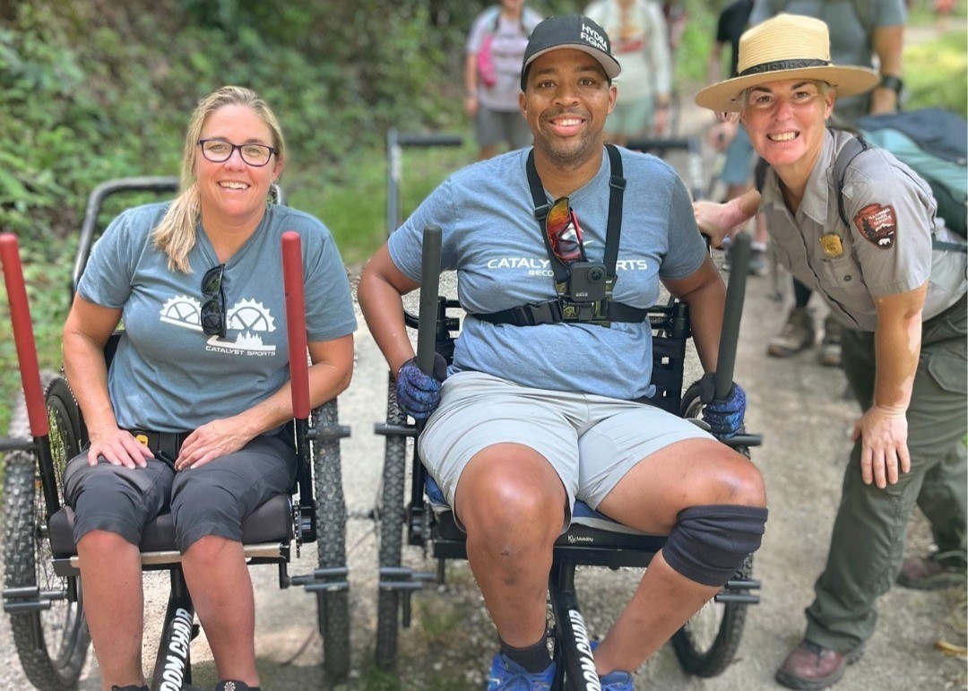 Two people in wheelchairs and a ranger pose for a picture on a trail.