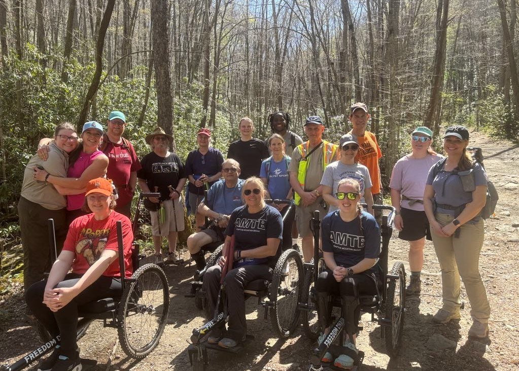 Large group of people, some in wheelchairs, pose for a picture on a forested trail.