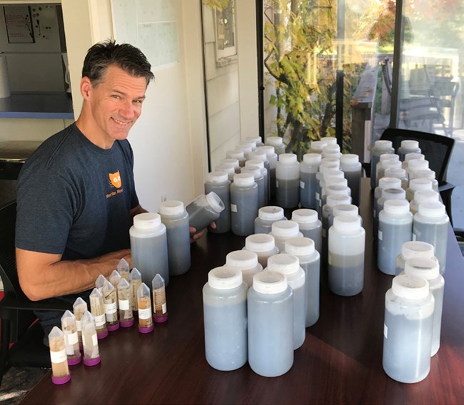 Smiling man sits at a table with several plastic liter water containers filled with dark liquid.