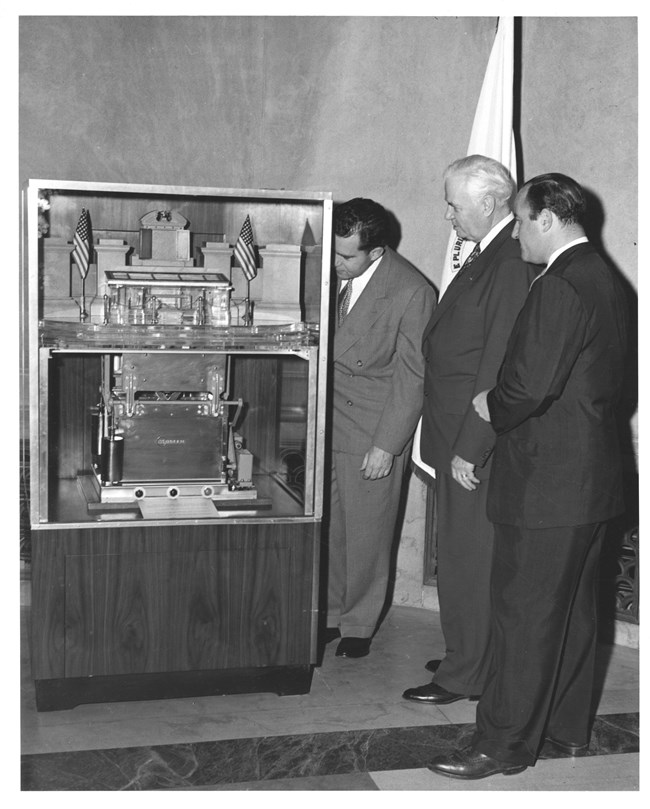 A model of the mosler safe is shown in a glass display case. Three men in suits are observing the model. The model shows the large safe below the viewing hall and shows how the Charters are moved between the safe and the rotunda.