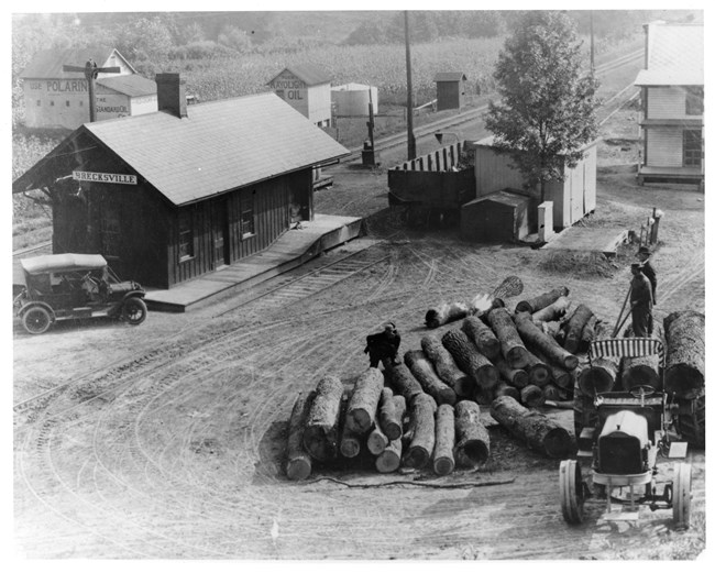 Four men stand on logs being unloaded from a truck with metal tires. A car is parked by a depot along railroad tracks with a cornfield beyond.