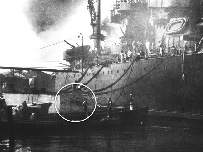 Black and white photo of metal battleship tied to cement mooring quay, cloud from explosion in background. In the foreground, mooring quay where “F6” is visible is circled to draw attention to it.