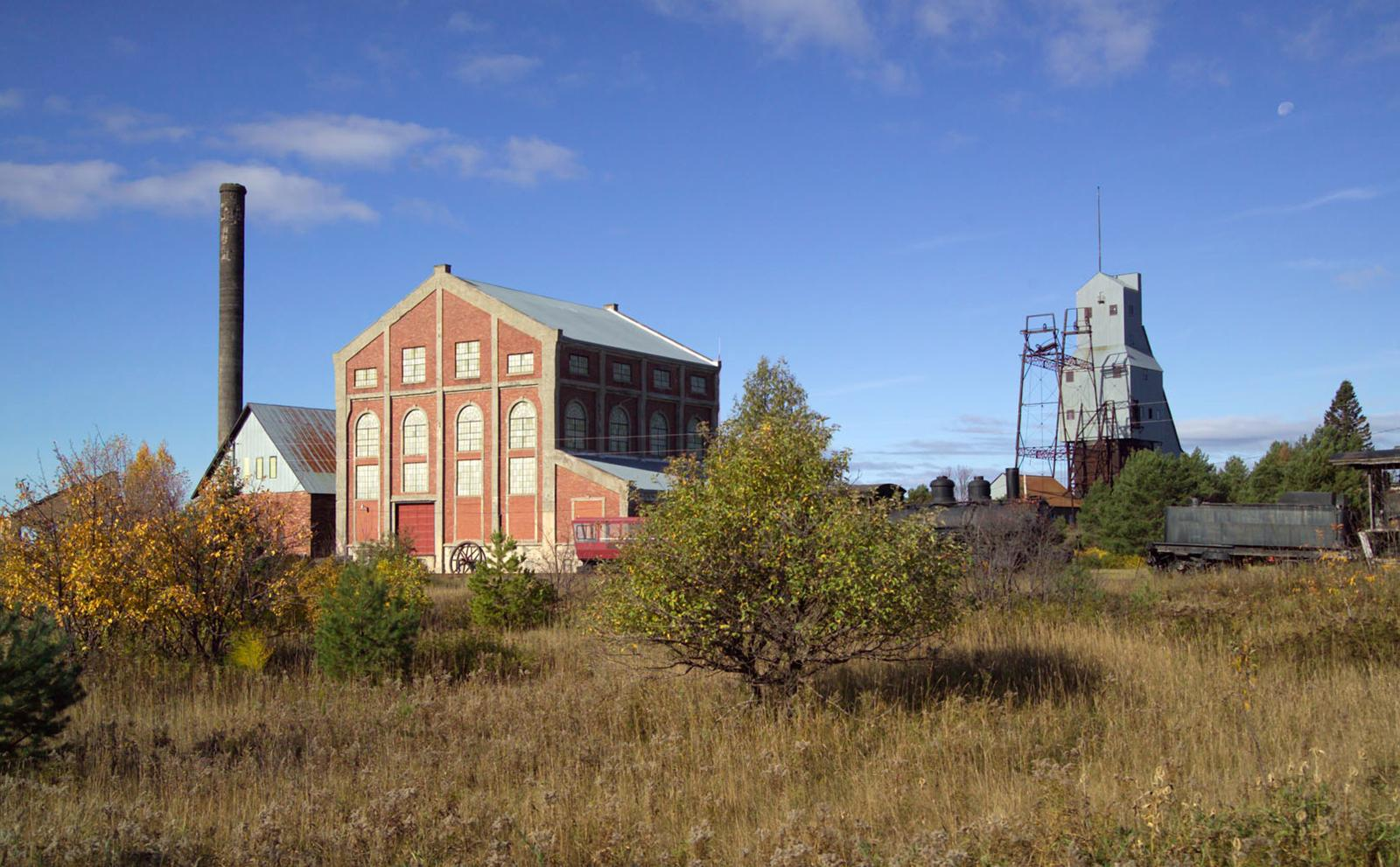 Photo of a large, red and grey industrial building. To the left is a smaller red building with a tall smokestack alongside it. To the right of these is a tall grey industrial building. Small trees and unkempt grass make up the foreground.