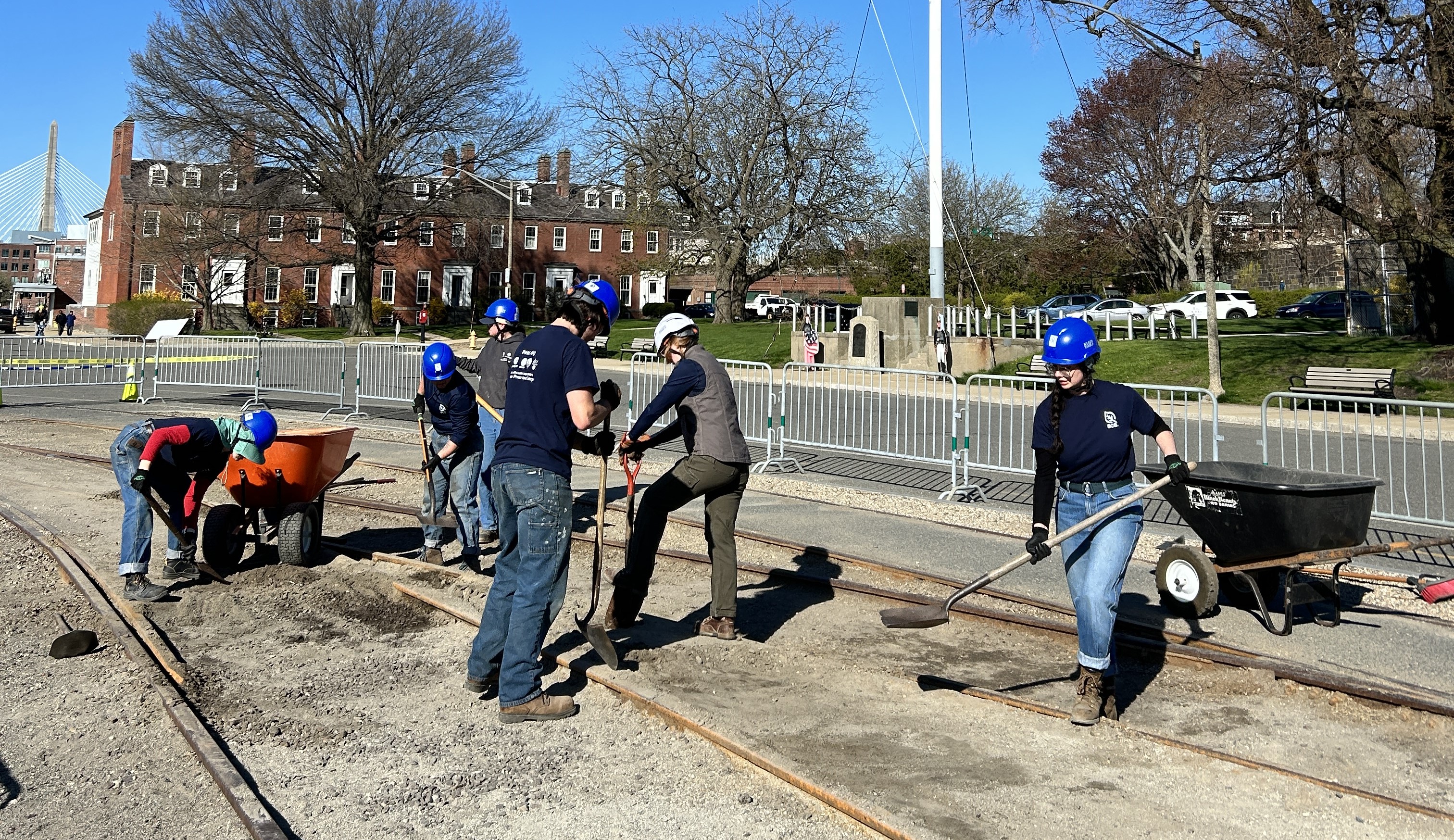 Six crew members with shovels, wearing hard hats, standing in an area of ground with gravel and trolley rails. They are shoveling the gravel out and putting it into two nearby wheelbarrows.
