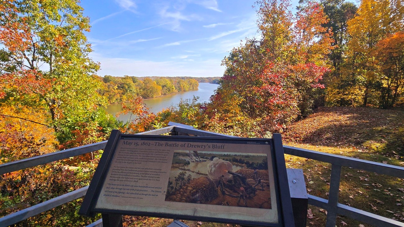 A sign titled "May 15, 1962 - The Battle of Drewry's Bluff" overlooking a tree-lined river on a beautiful fall day. The trees, from the foreground to the distance, are all a mix of green, orange, yellow, and bright red.