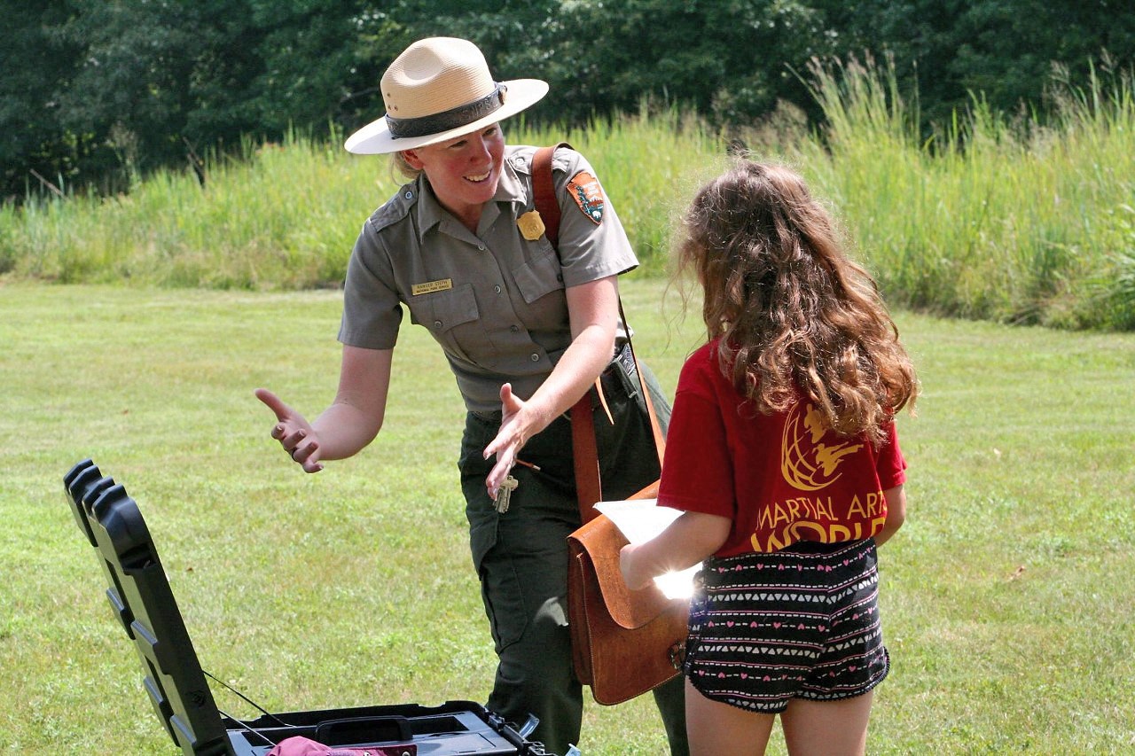 Woman outside in a green and gray uniform talking with a young girl and gesturing towards items in a black trunk.