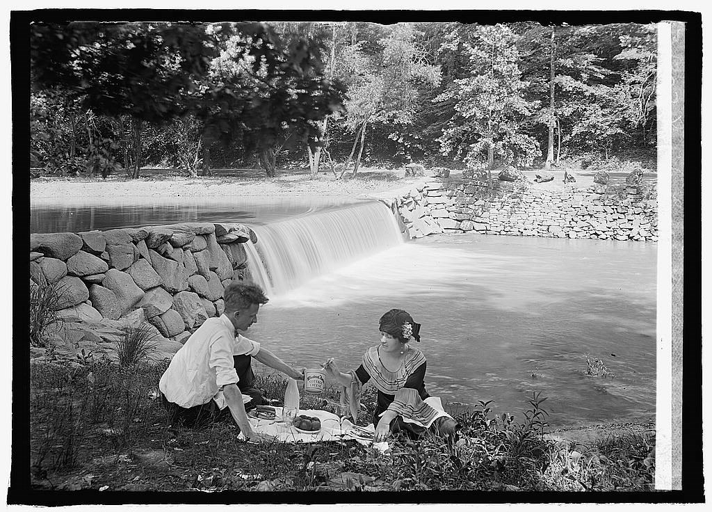 Two people sit on the ground with a picnic spread on a blanket between them, with a creek and waterfall in background. Black and white
