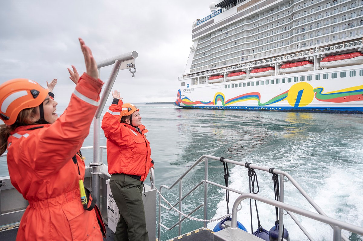 Two women in orange float coats and helmets wave to passengers on a passing cruise ship.