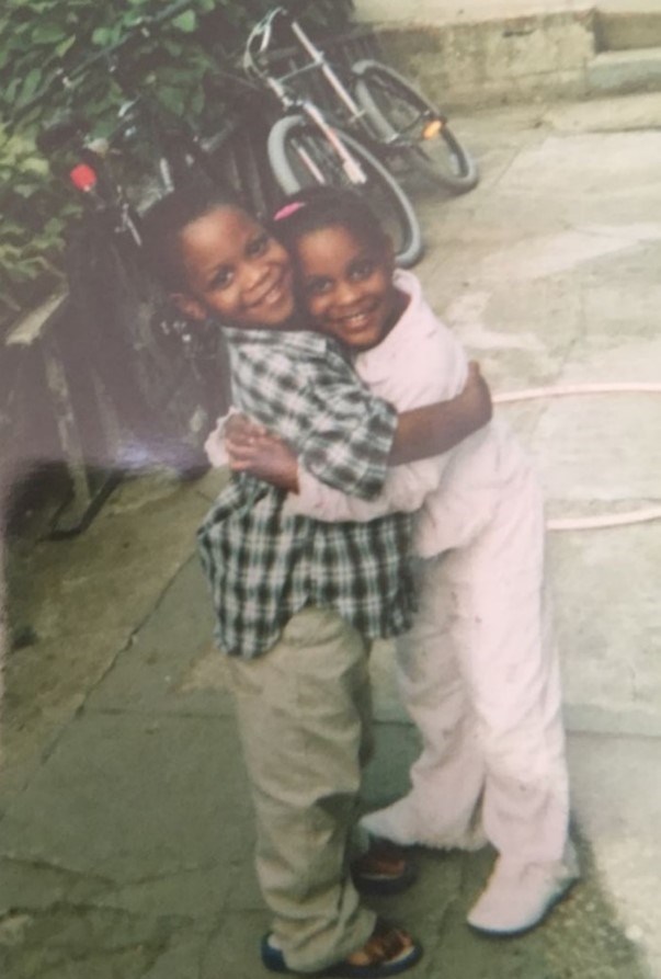 Two young siblings in an embrace while standing on a paved path with a bicycle and trees in the background to the left.