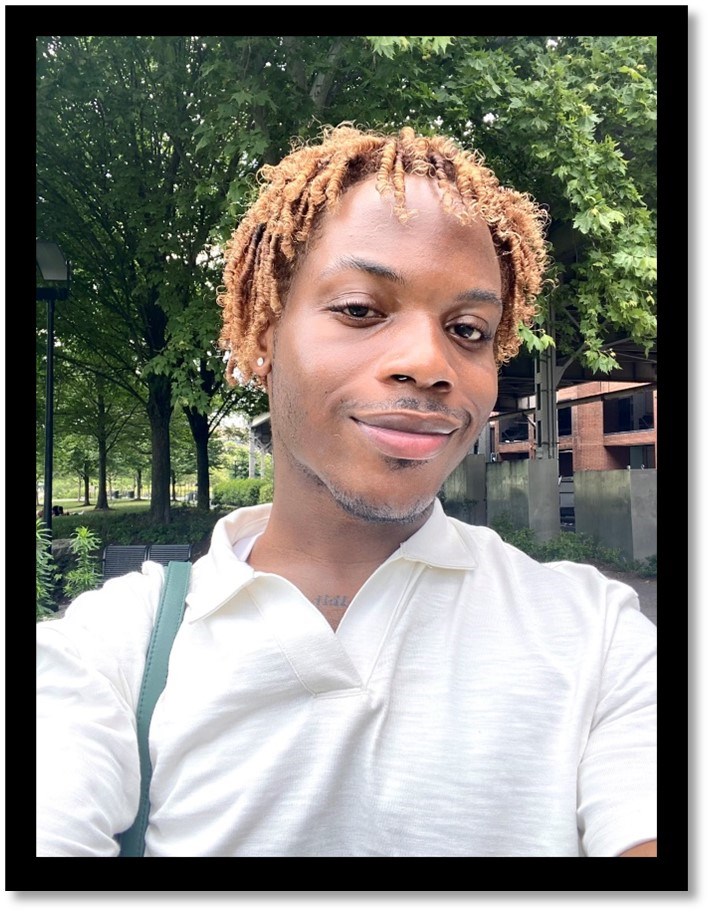 A young individual with short brown dreadlocks, brown shirt, and green trees in the background.