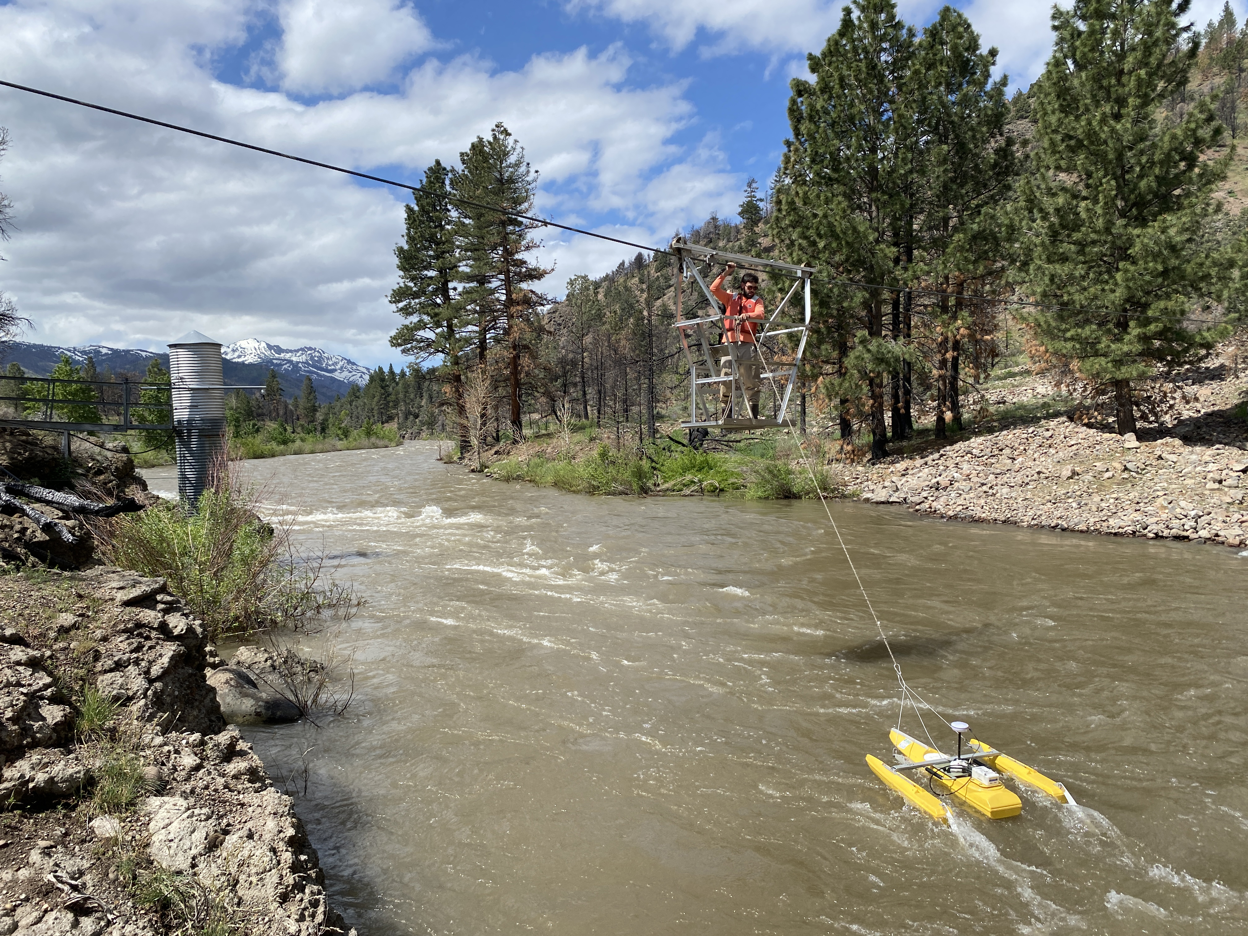 Person in a metal cage suspended from a cable over a river, holding on to a floating yellow flow measurement device via a long cable.