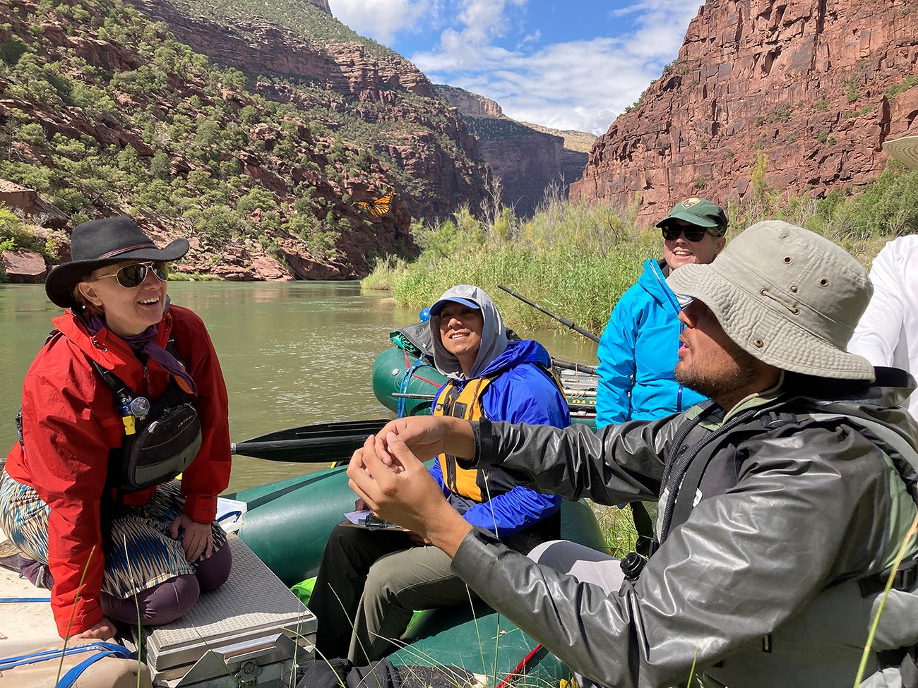 A group of people in a raft watch a Monarch fly away from them. A river and high bluffs are in the background.