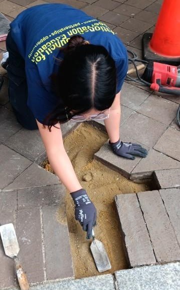 Woman with black hair, a blue shirt, and glasses. She is kneeling on bricks and holding a putty knife.