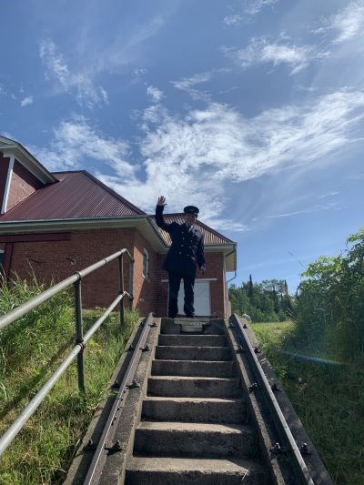 A man dressed in a black lighthouse keeper uniform waving outside from the top of concrete steps.