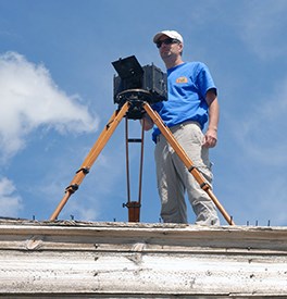 A man stands at a large camera on a tripod; blue skies in the background.