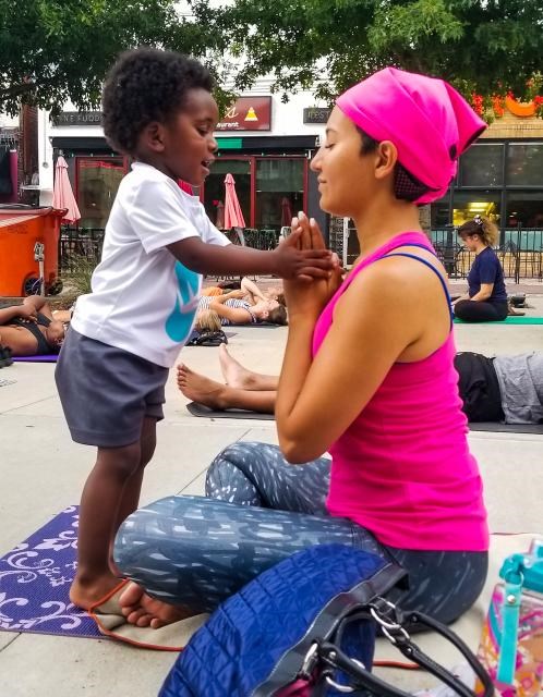 Riana Anderson sitting cross-legged on a yoga mat while a small child holds her hands together in a pose Riana Anderson sitting cross-legged on a yoga mat while a small child holds her hands together in a pose