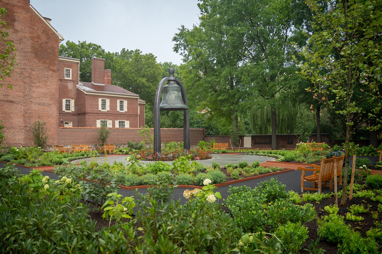 Tree and flowers surrounding gravel paths and a large bell in the middle.