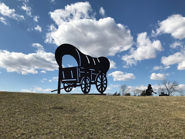 A silhouette of a covered wagon, on a grassy hill under a blue sky.