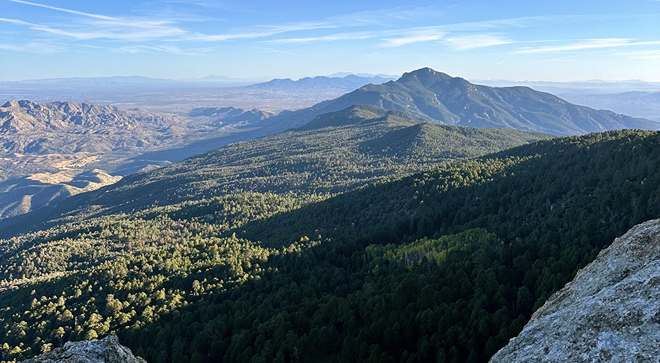 Looking down on magnificent conifer covered mountains, rugged desert hills, and desert valleys sprinkled with shadows under wispy clouds.
