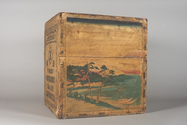 Wooden tea chest covered in brown paper showing a Japanese harbor with Mount Fuji in the background.