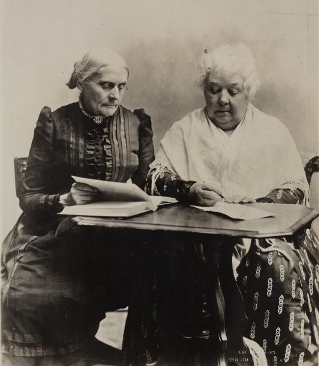 Two women, one seated, one standing, look at books. LOC