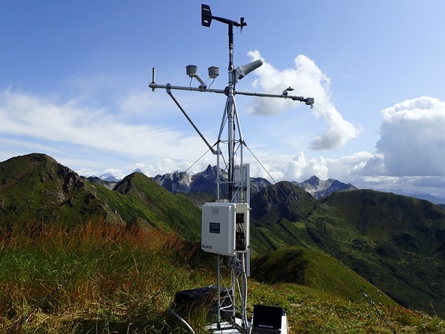 Weather station sits on a green hilltop with rugged mountains behind.