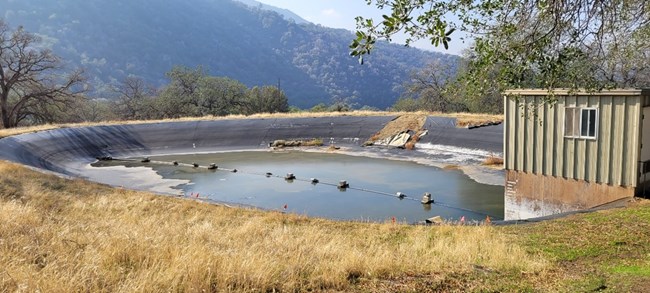 A light green shed sits beside a dilapidated looking man-made water basin with black lining. A mountain range is in the background.