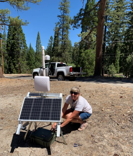 Ariane (Ari) Sarzotti, SEKI Smoke and Weather Technician, deploying a portable PA system with solar panel on level bare ground with white work truck and evergreen trees in background.
