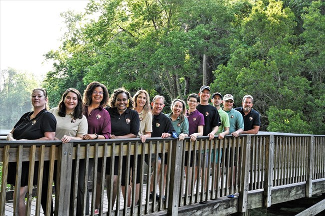 a group of eleven people, women and men, posing on a wooden bridge surrounded by nature
